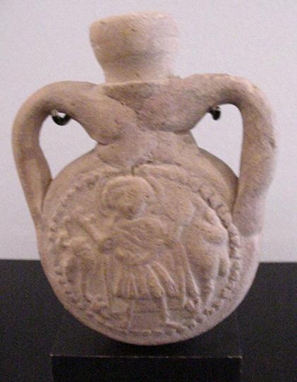 Pilgrims flask depicting St. Menas as an orant, flanked by two stylized kneeling camels. The reverse side contains the Blessing of St. Menas inscription within a wreath. Twin handles surround the center spout. These flasks were used to carry oil back from the shrine of the saint at Abu Mena, Egypt. A similar flask may be found at the Louvre. Front and back of flask with sharp images; one handle and part of neck restored, one handle original. Ca 6th century A.D. - 4" (10.1 cm. high)